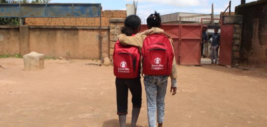 To skolepiger går mod skolen med armen om hinanden og nye røde Red Barnet skoletasker på ryggen